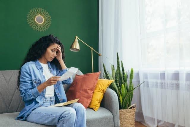 A woman in a blue denim outfit sits on a gray sofa in a room with a green wall looking concerned while reading a letter