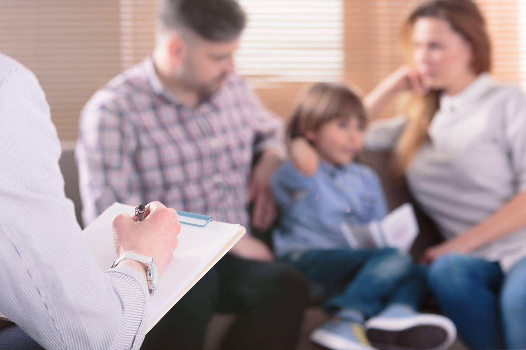 A man sits on a couch with his family writing on a clipboard as they enjoy each other's company