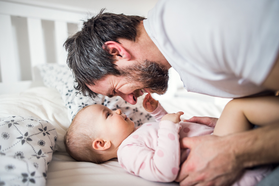 A man and a baby joyfully playing together on a bed, showcasing a moment of family bonding. Burton Family Attorneys.