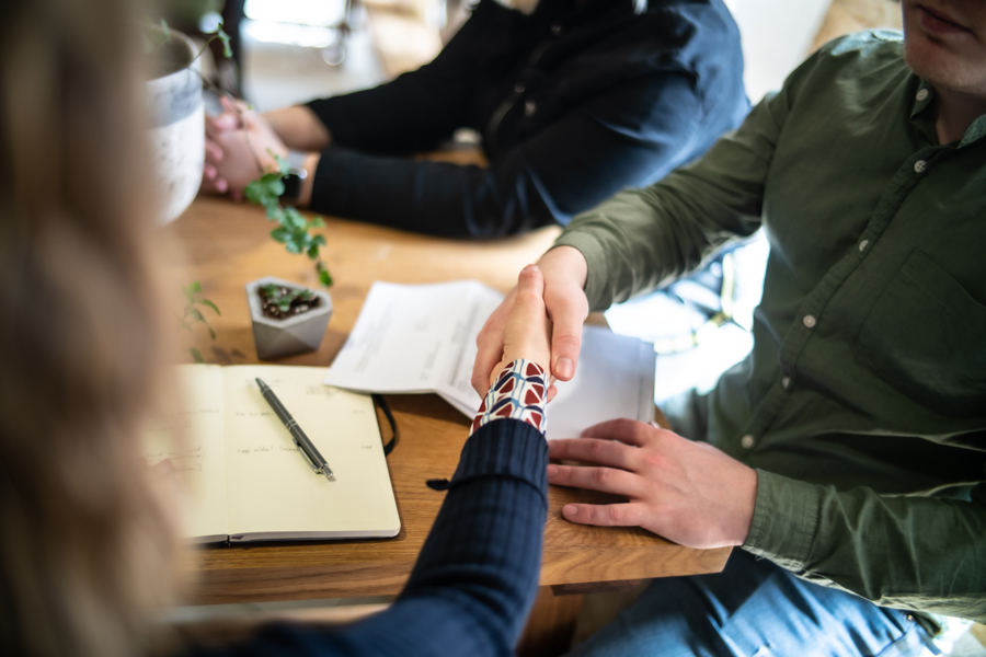 A handshake between two people across a wooden table in a casual office or meeting setting, with a notebook, pen, and documents visible, symbolizing agreement or collaboration.
