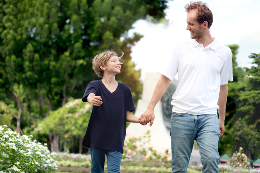A father and son walking hand in hand through a scenic park, smiling warmly at each other against a backdrop of green trees and blooming flowers.