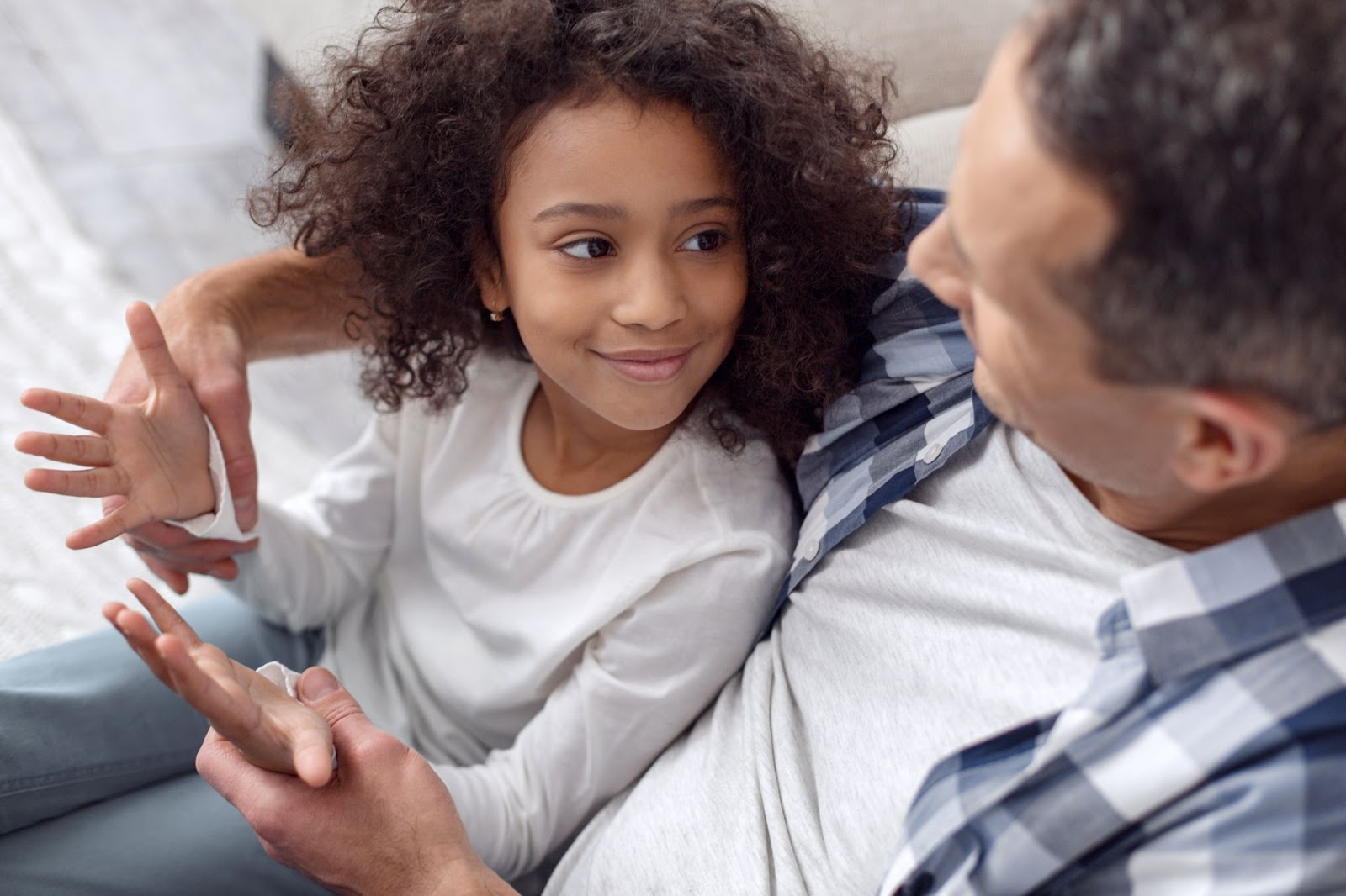 A child in a white shirt sits on an adult's lap, both in casual clothing, on a couch with a light-colored background.