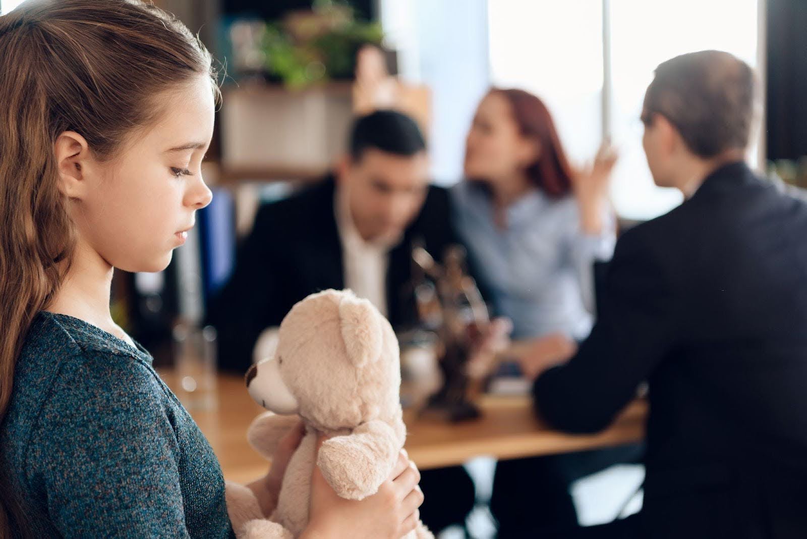 A little girl holds a teddy bear standing in front of a group of people who are smiling and watching her