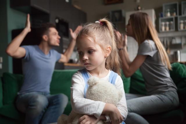 A little girl holds a teddy bear smiling in front of her family who stands together in a warm loving pose