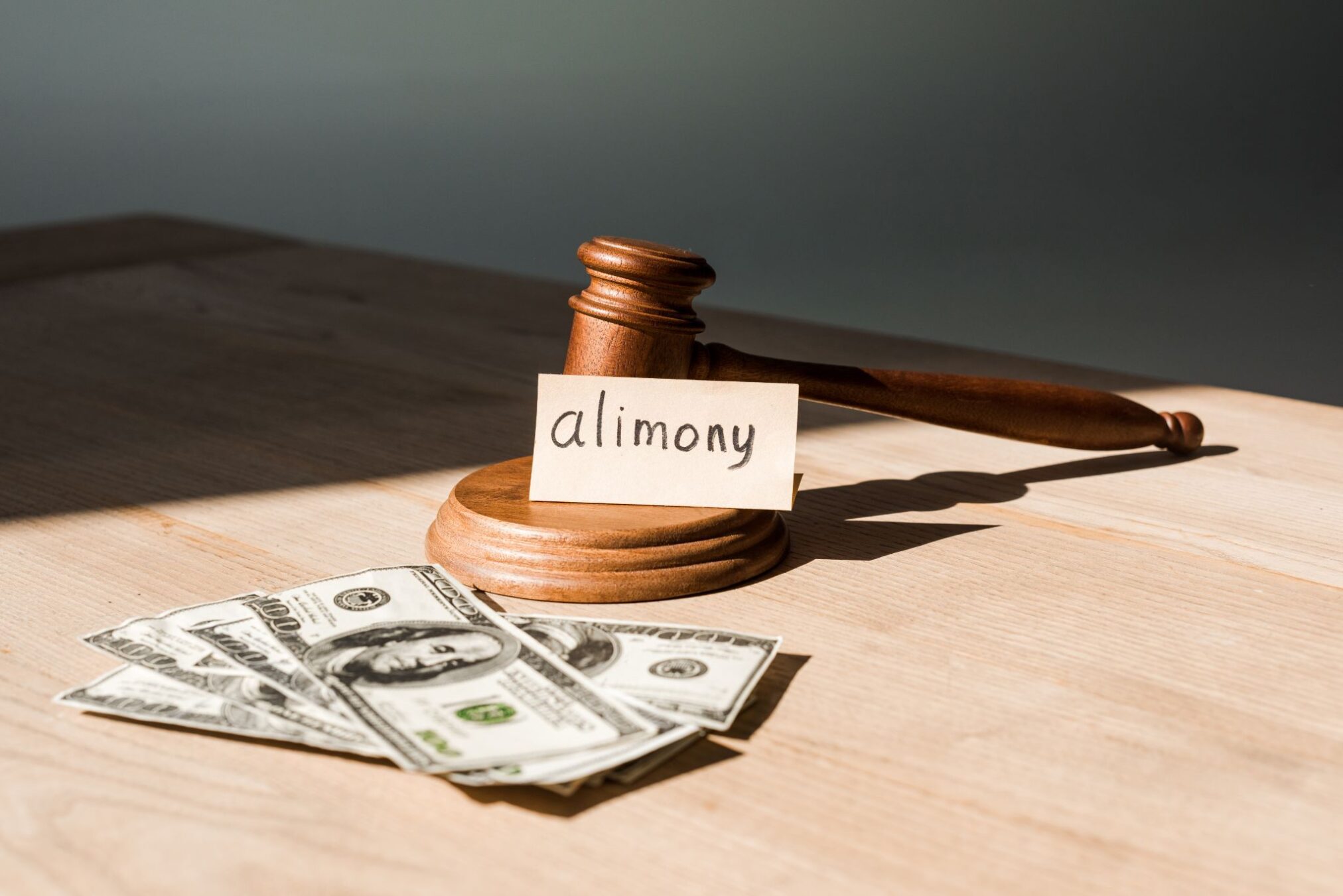 A wooden judge's gavel with an alimony card propped against it next to a fan of hundred-dollar bills.