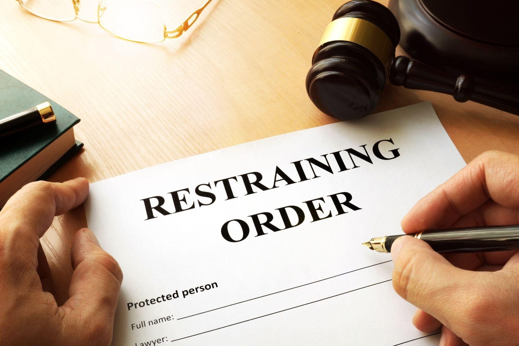 A person holds a pen poised to fill out a Restraining Order document on a wooden desk