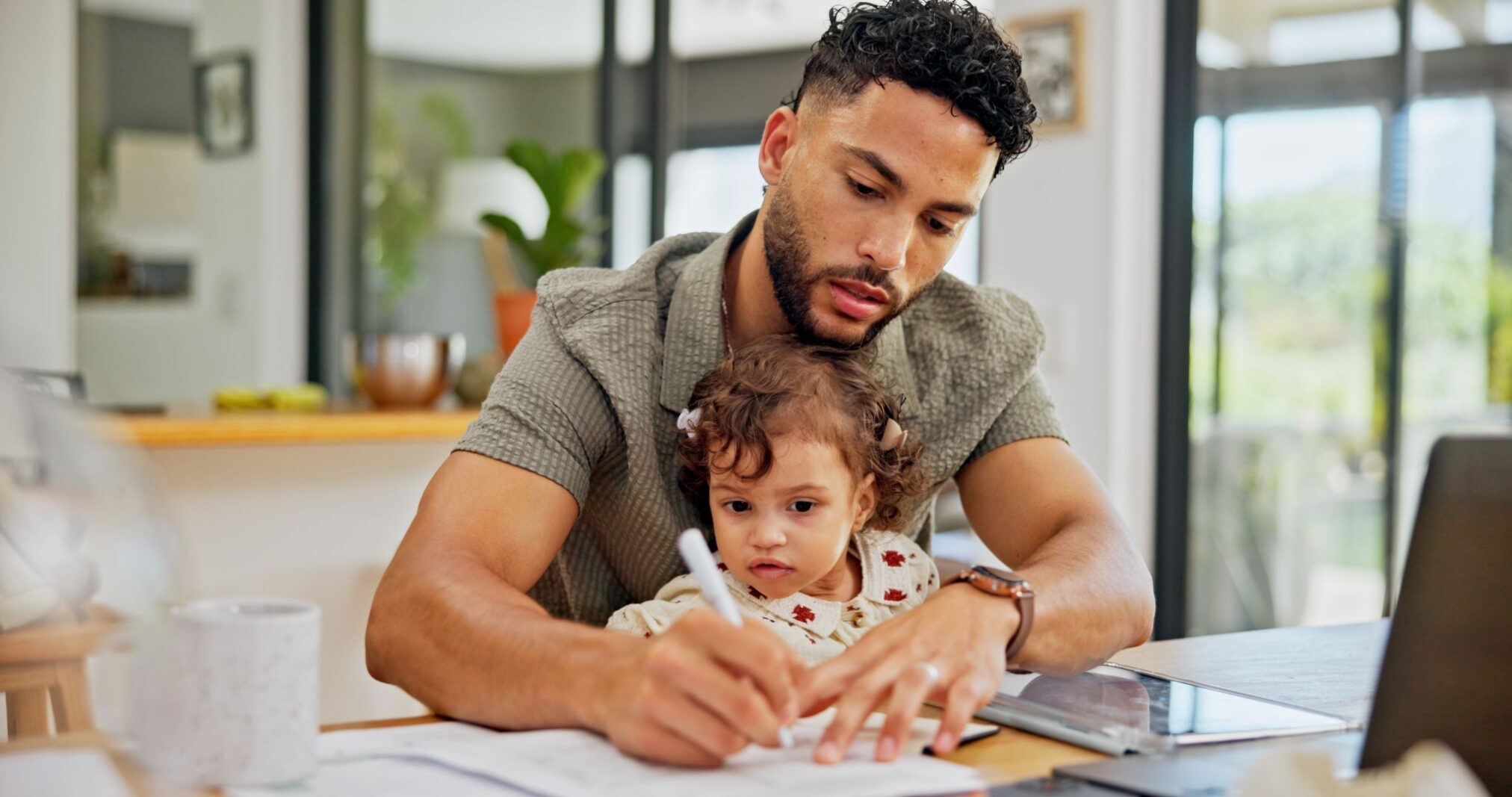 A father holding his toddler daughter while writing on paperwork at a desk.