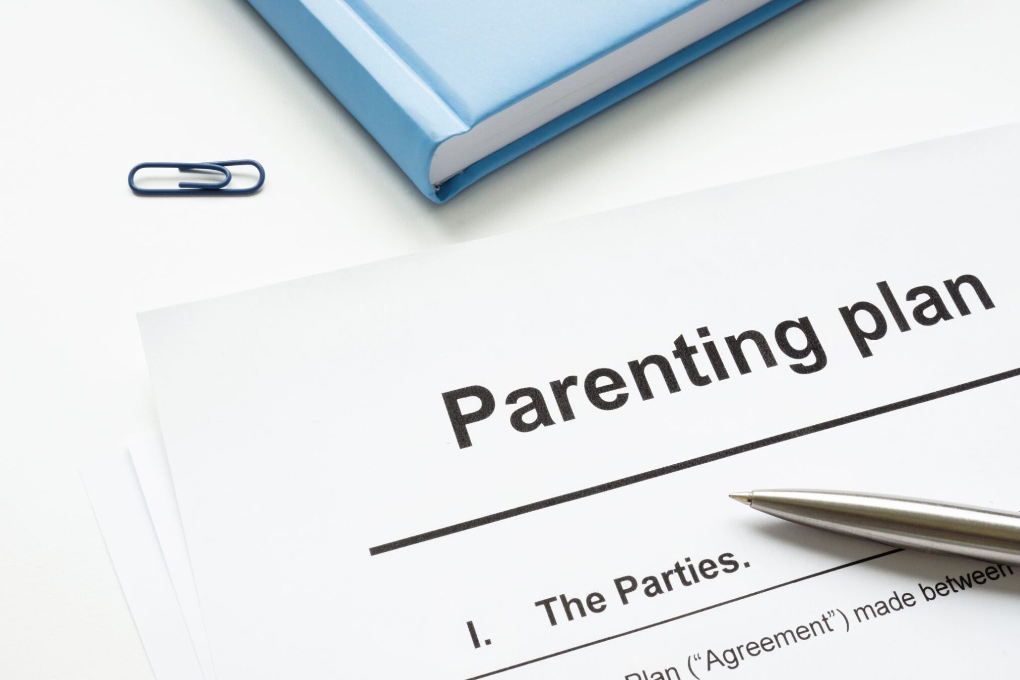 A legal document titled Parenting plan resting on a white desk next to a pen, blue book, and paperclip.